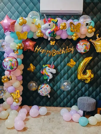 A unicorn-themed wall decoration for a 4th birthday. This setup includes a balloon garland, multiple unicorn and rainbow foil balloons, and a gold "Happy Birthday" banner against a dark, textured wall.