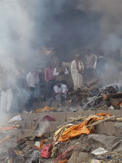 A wide shot of the cremation ghats, enveloped in smoke. This captures the scale of the rituals during Pitrpaksh, with a stray dog resting nearby, indifferent to the human activity.