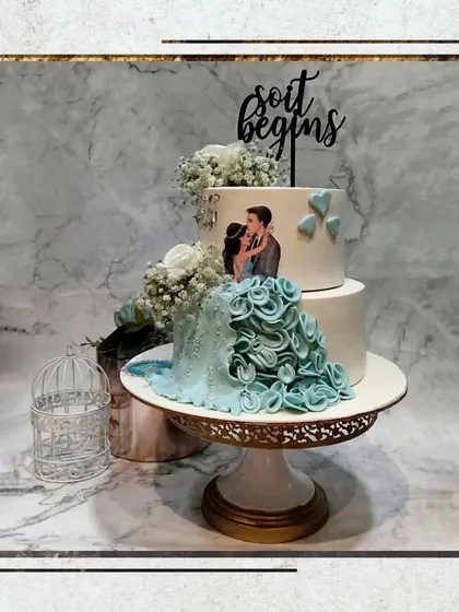 A two-tier engagement cake featuring a custom illustration of the couple and a beautiful ruffled dress detail. The "So it begins" topper perfectly captures the sentiment of the day.