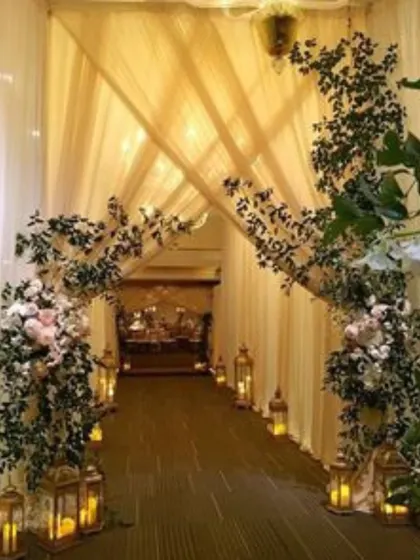 A rustic and romantic entranceway with an arch made of greenery and white flowers. The path is lit by lanterns, creating a cozy and inviting atmosphere for an evening celebration.