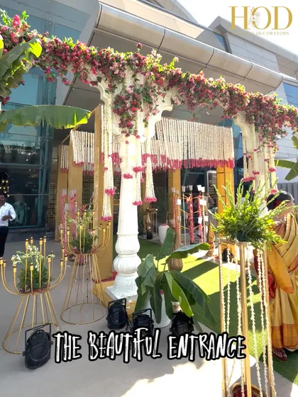 This outdoor wedding entrance combines traditional pillars with a canopy of bougainvillea and cascading white floral strings. The use of banana plants and golden candelabras adds a touch of South Indian tradition and opulence.