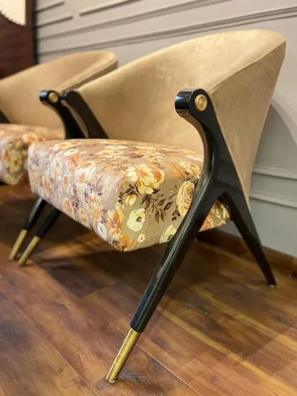 A side profile of a custom-designed accent chair, showcasing its unique silhouette. The chair combines a floral seat with a solid beige back, supported by an angular black wooden frame with brass-tipped legs.