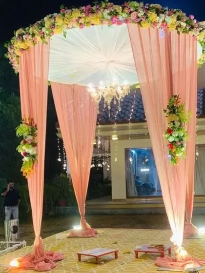 A beautiful and romantic outdoor wedding mandap. The design features a circular structure with delicate peach-colored drapes, a classic chandelier for evening ceremonies, and lush floral arrangements, creating a perfect setting for your vows.