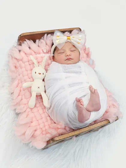 This little girl looks angelic in a simple white swaddle and a big bow headband, resting on a soft pink blanket with her bunny friend.