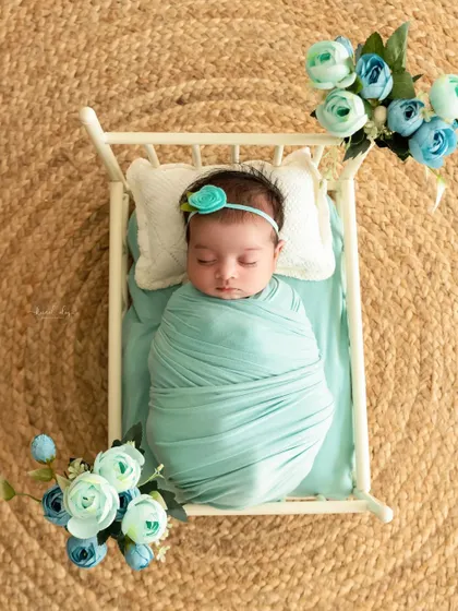 A newborn wrapped in mint green fabric sleeps in a miniature white crib, surrounded by matching blue flowers for a coordinated and peaceful scene.