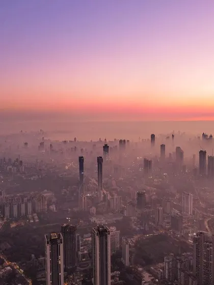 A wide drone shot of Mumbai enveloped in a sea of smog, with the city's skyscrapers barely visible through the haze at sunset.