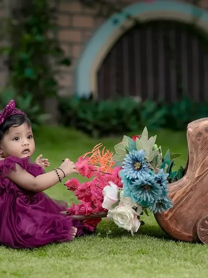 A baby girl in a beautiful purple dress, playing with a cart of flowers. Our garden is full of color and life.
