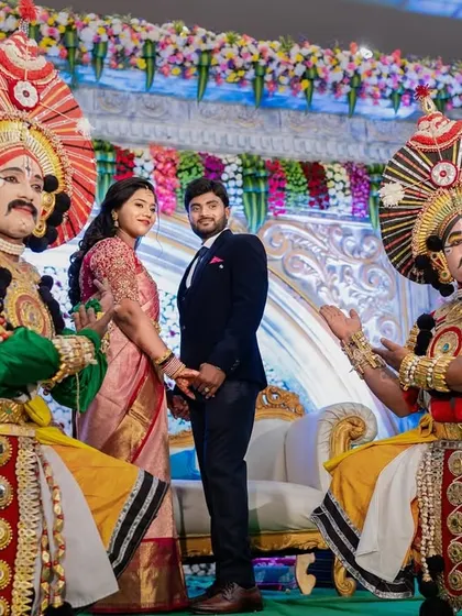 The couple stands with vibrantly dressed Yakshagana performers, adding a touch of traditional art to their modern reception.