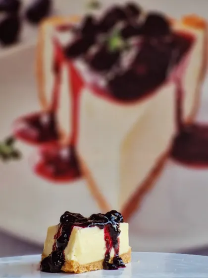 A creative shot capturing the essence of our Blueberry Cheesecake. A perfect mini bite in the foreground with the full slice artfully blurred in the back.