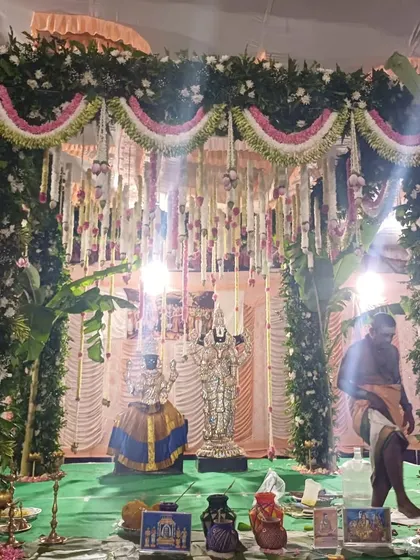 A traditional wedding ceremony in progress, with deities placed within the green and floral mandap.