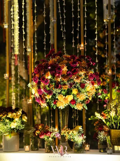 A large, vibrant bouquet of yellow, orange, and deep red chrysanthemums, set against a backdrop of hanging floral strings and candles.