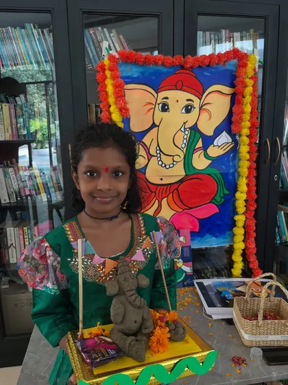 Dressed in festive attire, this student proudly presents her handmade Ganesha idol in front of a colorful backdrop.