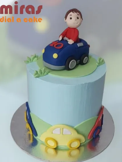 A sweet and simple car-themed cake perfect for a little one's birthday. A cute, blue fondant car sits atop a light blue cake, with more colorful cars decorating the sides for a playful touch.