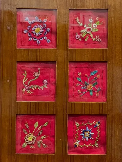 A set of six small, framed floral embroideries on a vibrant red background. These pieces are available for purchase and would look lovely as a collection in any room.