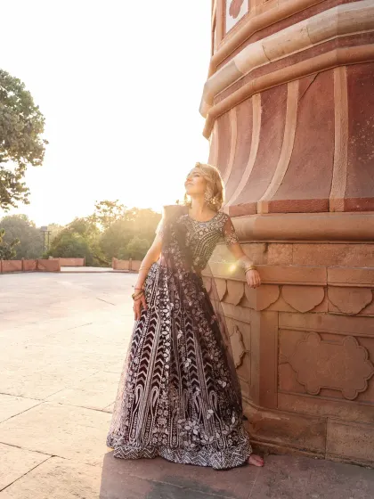A full-length shot of the model leaning against a historic sandstone pillar. The warm sunlight and her upward gaze create a feeling of serenity and grace, showcasing the full splendor of the lehenga.