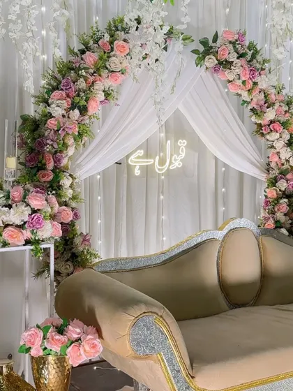 An elegant and serene Nikah backdrop. This setup features a beautiful floral arch in shades of pink and white, with delicate white drapes and a glowing 'Qubool Hai' neon sign, creating the perfect atmosphere for the ceremony.