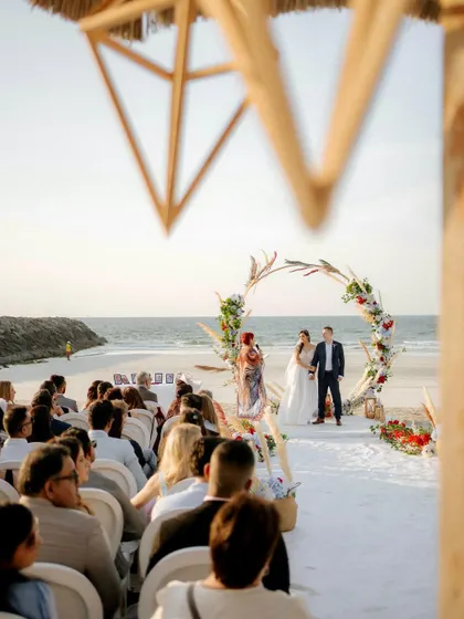 A view of the beach ceremony from the guests' perspective, showing the couple under a beautiful floral arch against the sea.