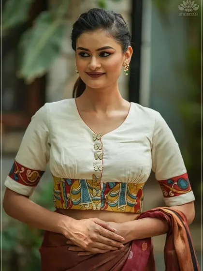 An elegant off-white blouse with a unique design. The waistband and sleeve cuffs feature a colorful Kalamkari panel, complemented by delicate butterfly-shaped buttons.