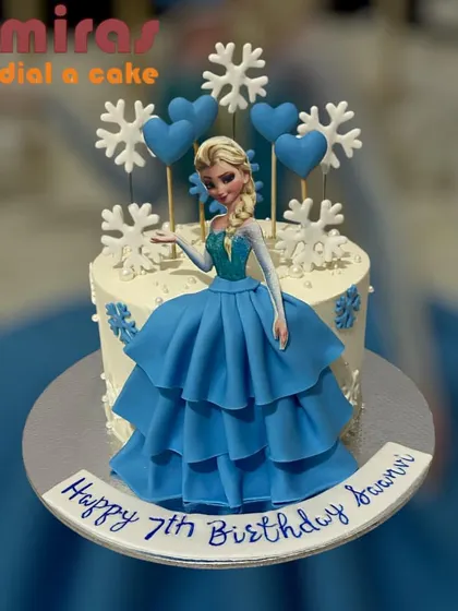 An elegant Elsa doll cake where the doll's dress is made of beautifully piped blue frosting. Snowflake and heart details add a touch of love to this magical Frozen-themed creation.