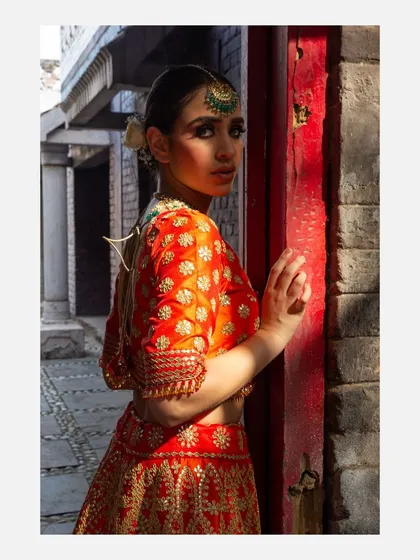 A model in a vibrant orange lehenga, captured in a narrow alley with strong directional sunlight. The play of light and shadow adds depth and drama to this fashion portrait.
