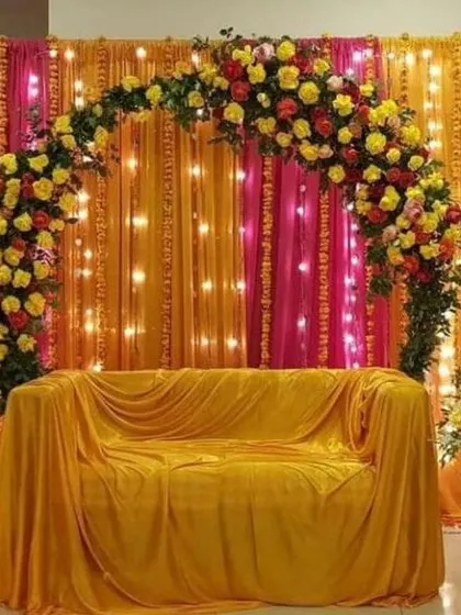A beautiful photo corner for a Haldi or Mehandi event, featuring a floral arch with yellow and red roses over a sofa covered in yellow fabric. The backdrop of pink and orange drapes with fairy lights adds a warm glow.