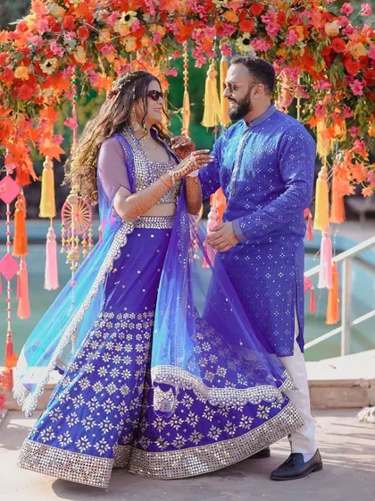 A playful moment between the couple at their Haldi ceremony. The bride's vibrant blue lehenga with mirror work stands out against the festive floral backdrop.