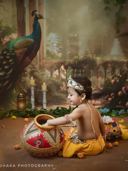 A classic Krishna pose, as this little boy reaches into the makhan pot. The artistic lighting and peacock backdrop create a painterly, timeless feel.