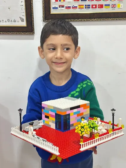 A young boy proudly holds his detailed architectural model of a house with a garden, built entirely from LEGOs. This project hones spatial reasoning and design skills.