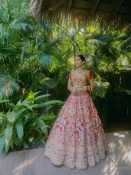 A full-length bridal portrait in a lush, tropical setting. The vibrant green of the foliage makes the bride's ornate red lehenga stand out beautifully.