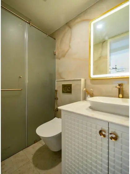 This bathroom has been transformed into a serene oasis with warm-toned marble walls, a vessel sink on a textured white vanity, and a gold-framed backlit mirror.
