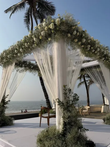A close-up of the beachside mandap, showing the delicate white drapes and the lush arch of baby's breath and white roses against the ocean backdrop.