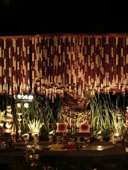 A wide view of the Banaras Ghat inspired mandap at night. The warm lighting highlights the dense floral ceiling, the rustic bell pillars, and the sacred elements, creating a deeply spiritual setting.
