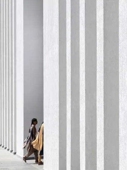 A person walks along a corridor at Sona University, framed by the strong vertical lines of the facade. This image captures a quiet moment of human experience within the larger architectural composition.