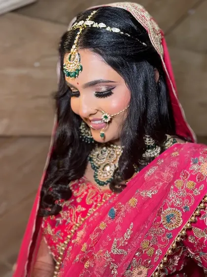 A smiling bride looking down, showcasing her beautiful soft-curl hairstyle and the intricate details of her pink lehenga.