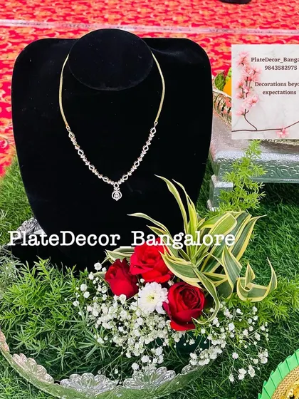 A simple gold necklace presented on a black bust, set within a glass platter decorated with fresh red roses and greenery.