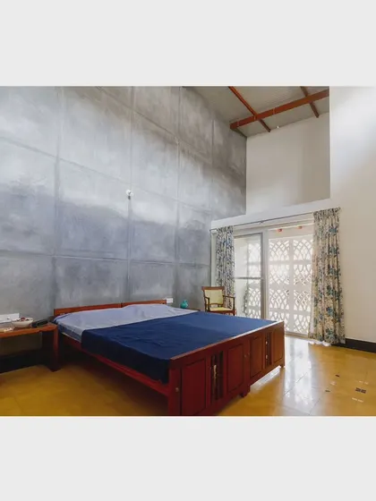 This bedroom in 'Malenadu Mane' has a dramatic double-height ceiling and a raw concrete accent wall. The design is minimalist, drawing attention to the volume of the space and the view outside.