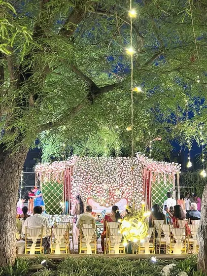 A wide view of an outdoor engagement ceremony at dusk, with trees lit by fairy lights and a beautiful floral stage.