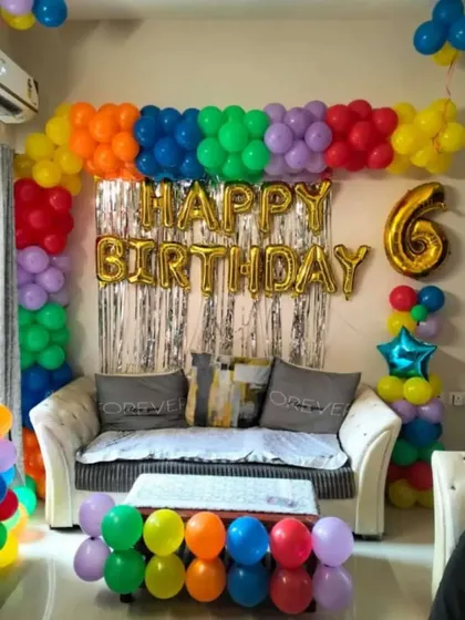 A vibrant rainbow-themed birthday decoration in a living room. The colorful balloon arch frames the sofa, making the whole space feel festive.