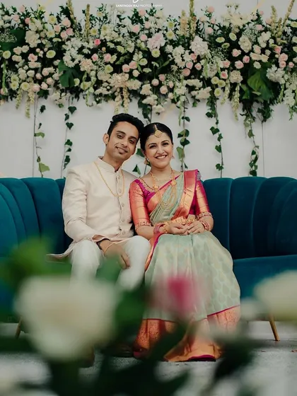 The couple seated against a stunning backdrop of white and blush flowers. The rich teal sofa provides a beautiful pop of color, making this a perfect photo corner at their engagement party.