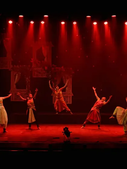 A wide shot of the ensemble on stage during "UNMASKED". The red lighting and expansive poses create a scene of high drama and energy, showcasing the scale of the production.