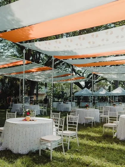 An elegant outdoor lunch setup with long tables under a canopy of orange and white drapes, providing shade and style for guests.