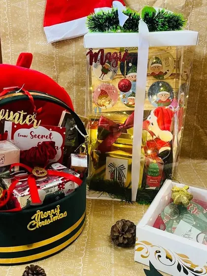 A festive collection of Christmas hampers. This display includes a large transparent bag with goodies, a round velvet 'Merry Christmas' box, and a smaller decorative gift box.