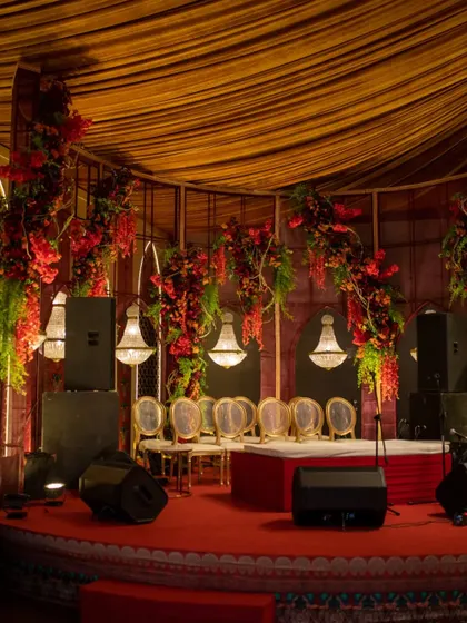 The stage area for a 'Mata ki Chowki', framed by dramatic arches of red flowers and greenery, with elegant chandeliers adding a divine glow. The design is both theatrical and reverent.