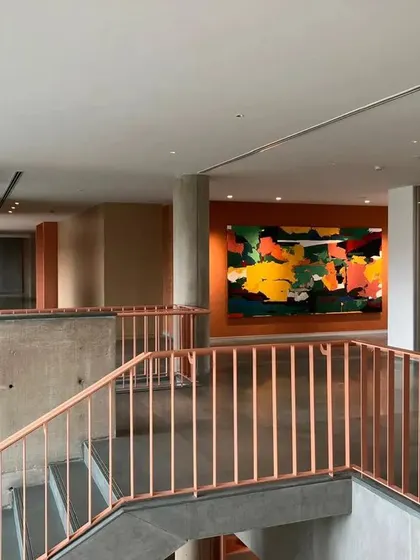 An interior view of a JSW office, where a colorful abstract painting provides a focal point in a circulation space. The warm tones of the railing and concrete walls create a welcoming and art-filled environment.