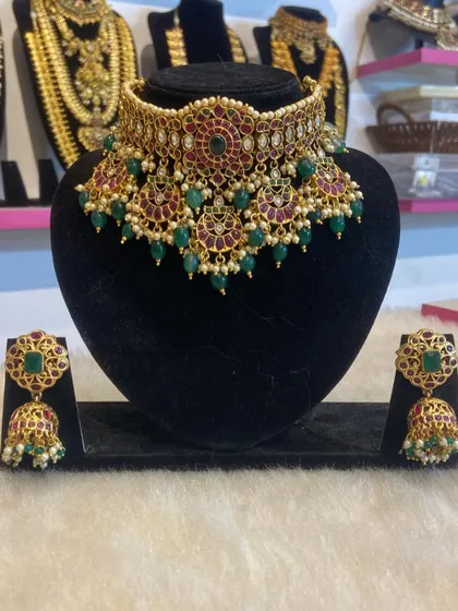 A stunning high-neck choker with ruby and emerald stones, paired with matching jhumkas. This is a perfect statement piece for a reception or engagement.