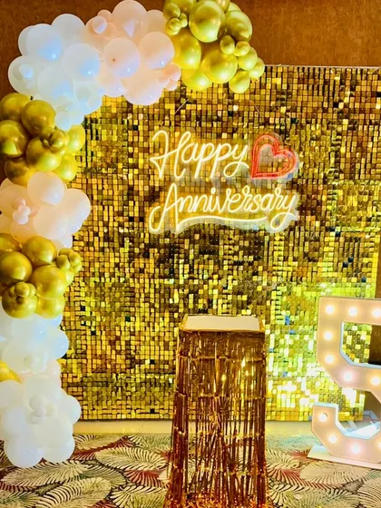 A classic 50th-anniversary setup with a gold shimmer wall and a white and gold balloon arch. The decor includes a "Happy Anniversary" neon sign and marquee numbers.