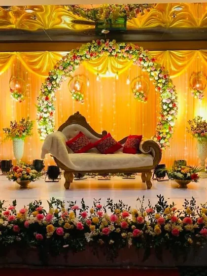 A warm and inviting reception stage with a full floral arch against a golden yellow backdrop. Hanging lanterns and a classic chaise lounge complete this traditional yet elegant look.