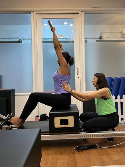 An instructor guides a client through a seated exercise on the reformer, offering support to ensure correct posture and muscle engagement.