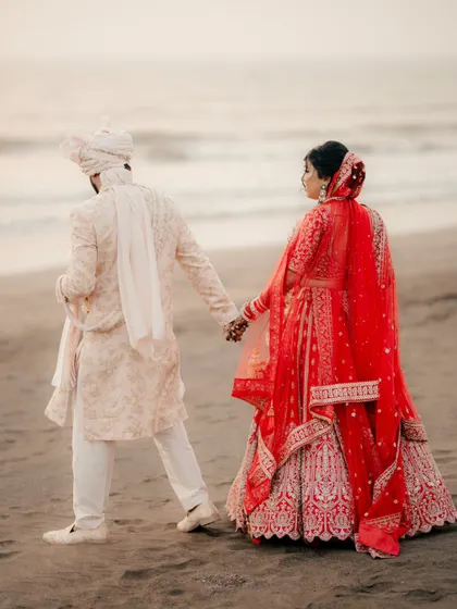 A walk to remember along the beach. This shot captures the couple hand in hand after their ceremony, with the bride's red lehenga vibrant against the sand.