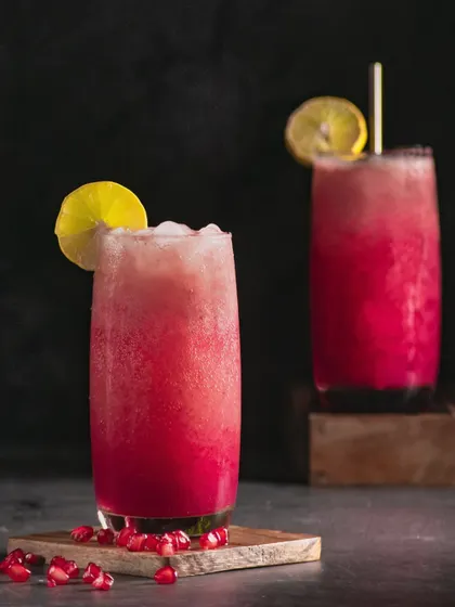 A pomegranate cooler with a lemon garnish. The dramatic lighting from behind makes the drink glow, emphasizing its vibrant pink color and fizzy texture.